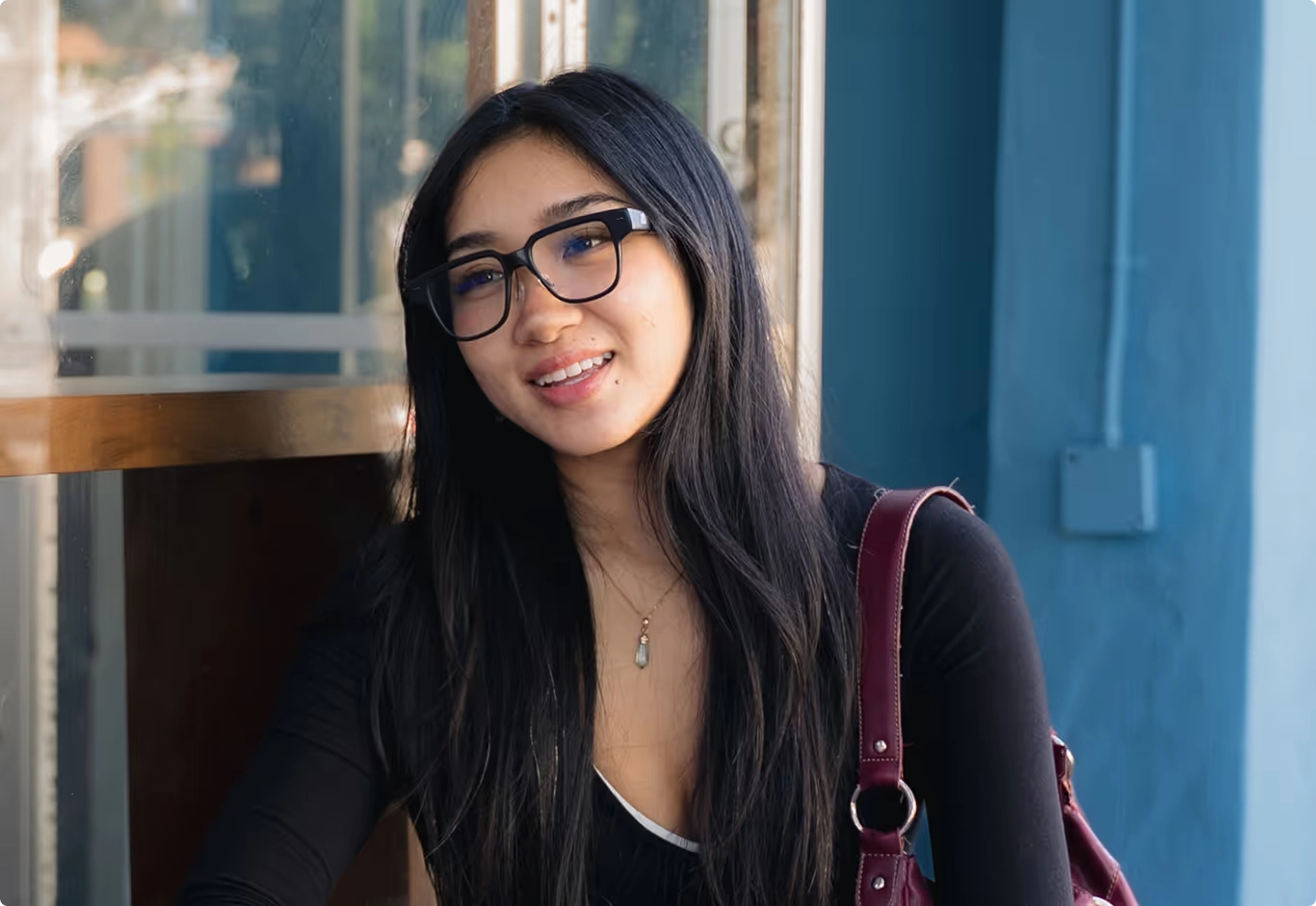 A smiling woman in black glasses standing by a blue wall